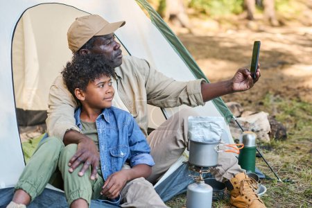 Téléchargez les photos : Homme afro-américain senior avec smartphone faire selfie avec son petit-fils tout en étant assis dans la tente et en regardant dans la caméra smartphone - en image libre de droit