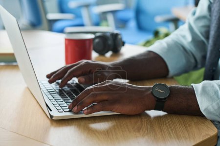 Foto de Manos del joven empresario afroamericano escribiendo en el teclado del ordenador portátil mientras está sentado en el lugar de trabajo en la oficina y en busca de datos en línea - Imagen libre de derechos