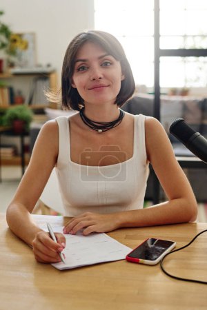 Photo for Young smiling brunette woman in casualwear sitting by workplace in front of camera and making notes while preparing for interview - Royalty Free Image