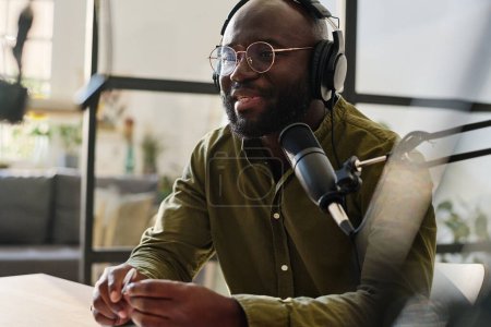 Foto de Joven hombre negro en auriculares y anteojos hablando en micrófono mientras está sentado en el lugar de trabajo y la grabación de archivos de audio para su audiencia - Imagen libre de derechos