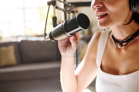 Photo for Close-up of young modern host in white tanktop speaking in microphone while sitting by workplace in front of camera during record - Royalty Free Image