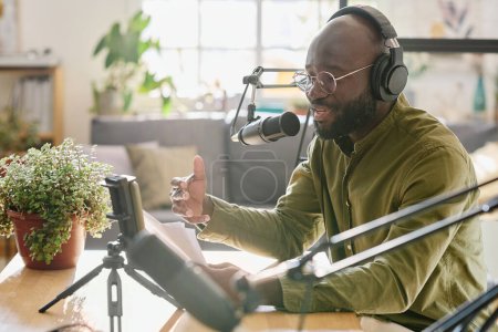 Téléchargez les photos : Black guy dans les écouteurs parler dans le microphone tout en lisant le plan de discussion et l'enregistrement de la nouvelle vidéo pour le public en ligne - en image libre de droit