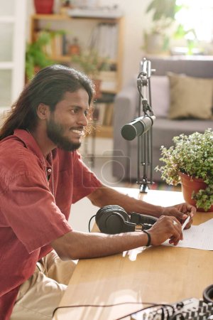 Téléchargez les photos : Jeune homme souriant aux cheveux longs foncés communiquant avec invité en studio assis à côté du bureau devant le microphone - en image libre de droit