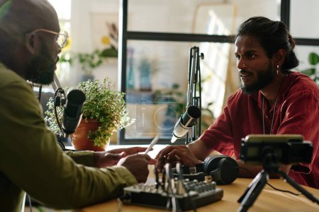 Téléchargez les photos : Jeune homme interculturel regardant le gars afro-américain lors d'une conversation par bureau dans un studio tout en parlant tous les deux dans des microphones - en image libre de droit