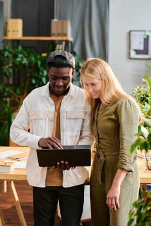 Photo for Two young intercultural employees in casualwear looking at laptop screen while standing by desk and preparing presentation of project - Royalty Free Image