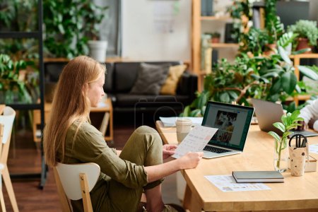 Foto de Joven empresaria creativa o diseñadora con documento mirando la pantalla del portátil con imágenes mientras está sentada en el lugar de trabajo en la oficina - Imagen libre de derechos
