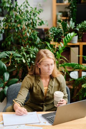 Téléchargez les photos : Jeune femme créative aux longs cheveux blonds prenant un café et prenant des notes dans le document tout en analysant les données en ligne sur écran d'ordinateur portable - en image libre de droit