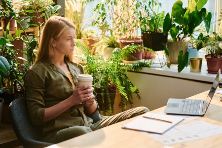 Téléchargez les photos : Jeune femme blonde avec une tasse de café regardant l'écran d'ordinateur portable pendant le réseau ou tout en regardant la vidéo en ligne ou en communiquant - en image libre de droit