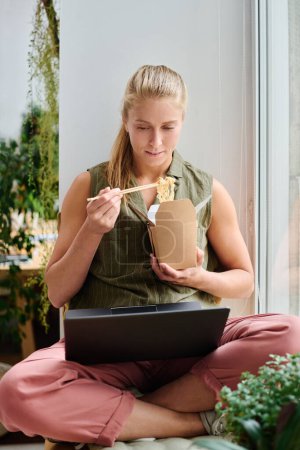 Téléchargez les photos : Jeune femme d'affaires affamée avec ordinateur portable sur ses jambes croisées manger wok chinois et regarder la vidéo en ligne tout en étant assis par la fenêtre - en image libre de droit