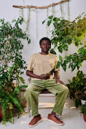 Téléchargez les photos : Jeune homme afro-américain en tenue décontractée avec smartphone assis sur une chaise parmi les plantes vertes au bureau et regardant la caméra - en image libre de droit