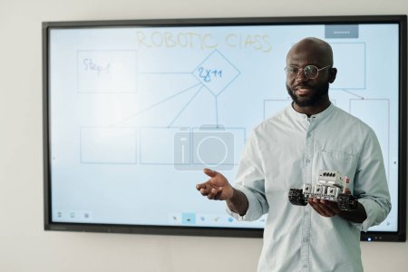 Téléchargez les photos : Jeune professeur de classe robotique tenant un nouveau modèle de robot jouet à la leçon tout en se tenant près du tableau blanc devant le public - en image libre de droit