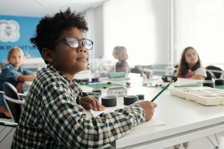 Foto de Escolar inteligente en anteojos y camisa a cuadros haciendo notas en copybook mientras está sentado junto a la mesa contra sus compañeros de clase - Imagen libre de derechos