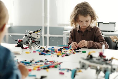 Photo for Clever schoolboy sitting by desk and connecting details of robot together while constructing new electronic toy at lesson of robotics - Royalty Free Image