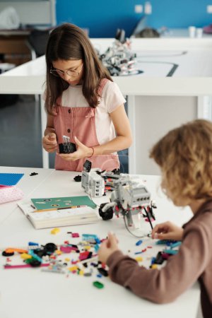 Photo for Two elementary schoolkids standing by table with details of robot at lesson of robotics and using manual while connecting them together - Royalty Free Image