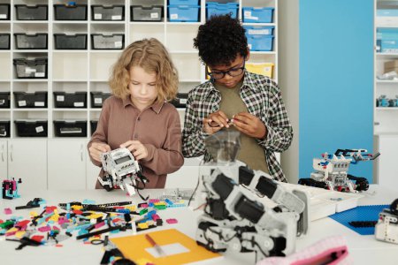 Photo for Two cute intercultural schoolboys connecting together details during construction of new robots at lesson of robotics at school - Royalty Free Image