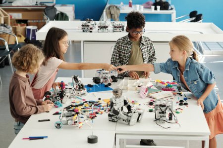 Photo for Group of youthful intercultural schoolkids constructing new robots at lesson while helpful schoolgirl passing detail to classmate - Royalty Free Image