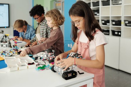 Foto de Fila de cuatro estudiantes interculturales de la escuela primaria que construyen nuevos robots de juguete mientras están de pie a la mesa con detalles en el aula - Imagen libre de derechos