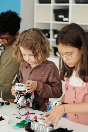 Foto de Grupo de escolares interculturales juveniles que conectan detalles mientras crean nuevos robots de juguete en la lección de robótica en la escuela primaria - Imagen libre de derechos