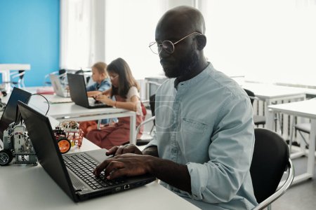 Téléchargez les photos : Sérieux jeune homme noir en chemise décontractée assis devant un ordinateur portable à la leçon en classe contre deux écolières diligentes réseautage par bureau - en image libre de droit