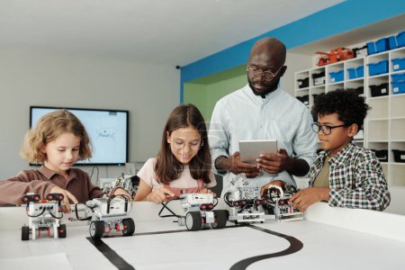 Foto de Joven profesor afroamericano confiado de robótica con tableta ayudando a escolares interculturales lindos a controlar sus robots durante el juego - Imagen libre de derechos