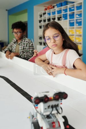 Photo for Clever youthful schoolgirl in eyeglasses looking at toy robot moving along table while standing against African American classmate - Royalty Free Image