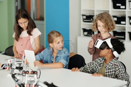 Téléchargez les photos : Écolière mignonne avec tablette regardant son camarade de classe dans un casque vr jouer à un jeu virtuel ou faire la présentation d'un nouveau robot à la leçon - en image libre de droit