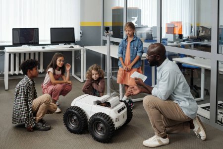 Foto de Joven profesor de robótica con tableta haciendo presentación de rastreador a grupo de escolares interculturales mientras controla robot de forma remota - Imagen libre de derechos