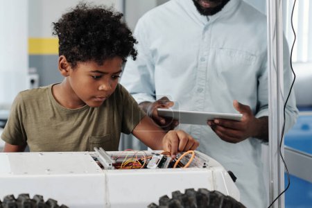 Foto de Lindo colegial afroamericano conectando cables de robot de juguete mientras sigue las instrucciones de su profesor de robótica con tableta - Imagen libre de derechos