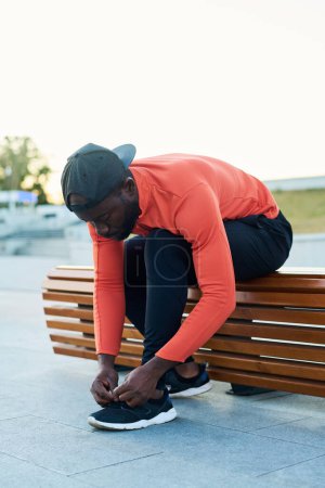Photo for Young sportsman in activewear tying shoelaces of sneakers while sitting on wooden bench in park or urban environment - Royalty Free Image