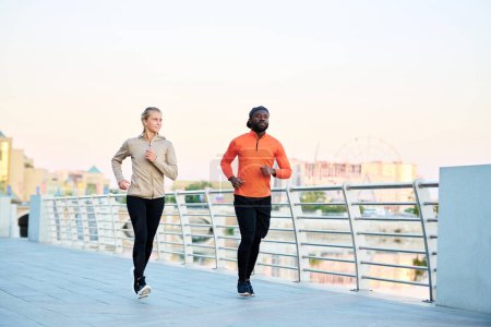 Photo for Two young intercultural athletes in sport jackets, leggins and sneakers jogging along bridge by riverside in the morning - Royalty Free Image