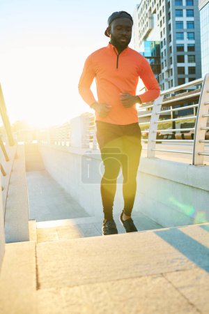 Foto de Joven deportista afroamericano corriendo por el puente en el entorno urbano por la mañana con la luz del sol detrás - Imagen libre de derechos