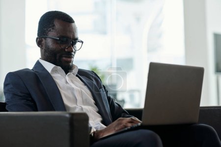 Foto de Joven empleado afroamericano serio con computadora portátil de rodillas en red mientras está sentado en un sillón contra el interior de la oficina - Imagen libre de derechos