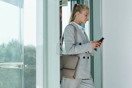 Foto de Serio joven rubia empleada mensajes de texto en el teléfono inteligente al salir del ascensor y salir del centro de la oficina para la hora del almuerzo - Imagen libre de derechos