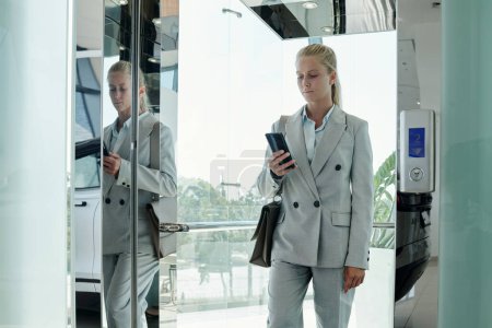 Foto de Joven mujer de negocios moderna en gris formalwear mensajes de texto en el teléfono móvil mientras sale del ascensor en el segundo piso del centro de oficina - Imagen libre de derechos