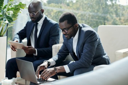 Téléchargez les photos : Jeune homme noir sérieux en formalwear dactylographier sur clavier d'ordinateur portable tout en étant assis dans le fauteuil contre collègue masculin en utilisant la tablette - en image libre de droit