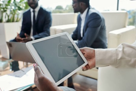 Téléchargez les photos : Tablette avec écran noir dans les mains de la jeune femme d'affaires afro-américaine réseautage contre deux collègues masculins - en image libre de droit