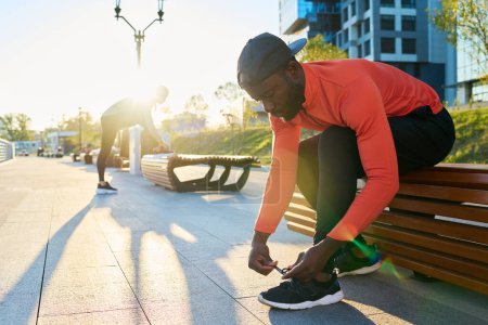 Téléchargez les photos : Jeune sportif afro-américain en veste de sport rouge et leggins noirs attachant lacet de chaussure de sneaker tout en se penchant vers l'avant sur la jambe - en image libre de droit
