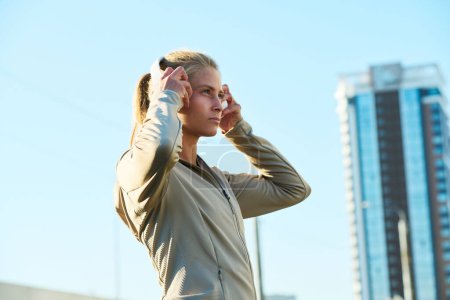 Téléchargez les photos : Vue latérale de la jeune femme blonde en veste de sport grise mettant des écouteurs sur sa tête tout en se tenant contre le ciel bleu et le gratte-ciel - en image libre de droit