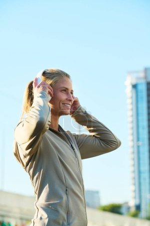 Téléchargez les photos : Vue latérale d'une jeune sportive souriante écoutant de la musique dans un casque tout en se tenant devant une caméra dans la ville - en image libre de droit