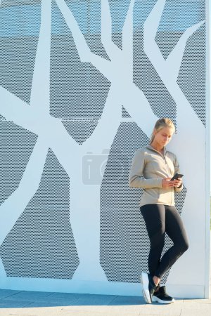 Foto de Joven deportista rubia en leggins negro y chaqueta deportiva gris de pie junto a la pared del edificio moderno y desplazándose a través de contactos en el teléfono - Imagen libre de derechos