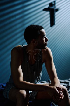 Téléchargez les photos : Jeune homme fatigué et malheureux regardant clair de lune venant par la fenêtre de sa chambre tout en étant assis sur le lit à minuit - en image libre de droit