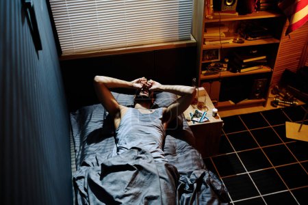 Téléchargez les photos : Jeune homme insomniaque avec syndrome post-traumatique gardant les mains sur la tête alors qu'il était couché et souffrant d'insomnie ou de maux de tête - en image libre de droit