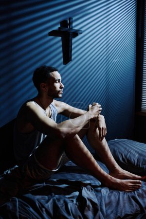 Photo for Young depressed man sitting on bed while staying awake all night long till morning and looking at window with venetian blinds - Royalty Free Image