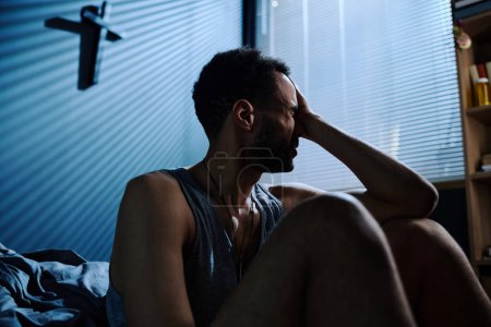 Photo for Young stressed man touching head while sitting by bed after sleepless night and feeling helpless about consequences of PTSD - Royalty Free Image