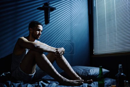 Photo for Young stressed man suffering from insomnia sitting on bed against wall with cross while morning light penetrating through venetian blinds - Royalty Free Image