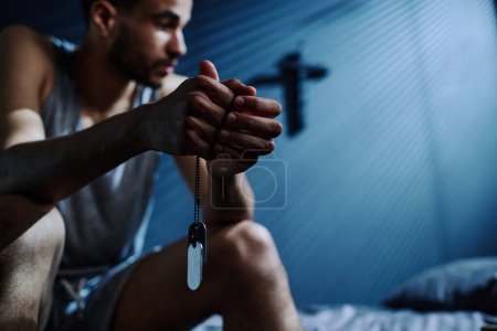 Foto de Manos de hombre joven insomne sosteniendo cadena de plata con medallones mientras está sentado en la cama después de la noche sin dormir y pensando en algo - Imagen libre de derechos