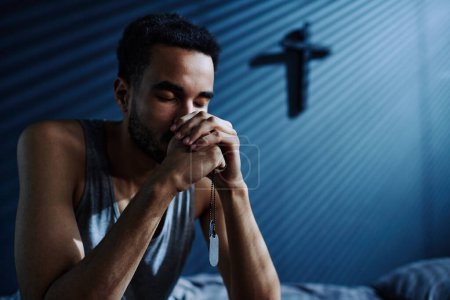 Photo for Young depressed man with metallic medallions on chain in his hands praying God to help him get asleep while sitting on bed - Royalty Free Image