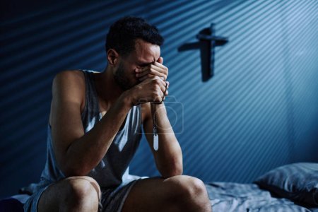 Photo for Young depressed man with two medallions on silver chain crying or grieving about loss of someone he loved while sitting on bed at night - Royalty Free Image