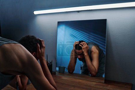 Foto de Joven con trastorno postraumático de pie frente al espejo con la cabeza en las manos mientras siente ansiedad e impotencia - Imagen libre de derechos