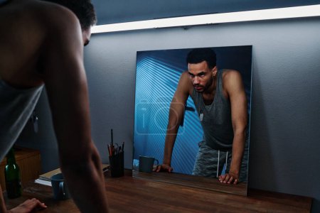 Photo for Young sleepless man with post traumatic syndrome looking at himself in mirror while bending over table in bedroom at night - Royalty Free Image